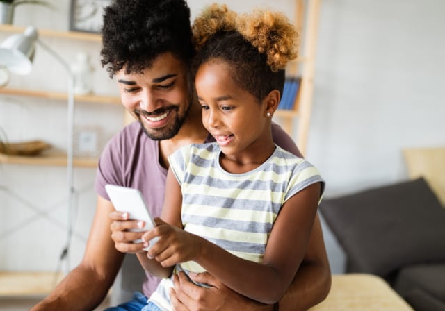Young daughter sitting on her father’s lap while learning how to use a new cell phone.